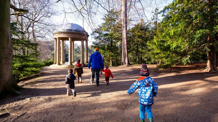 A family dressed in coats and hats walking along a path in the parkland at Fountains Abbey and Studley Royal Water Garden, surrounded by trees with a small historic structure in the background